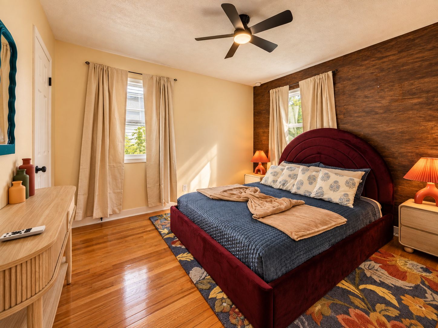 Un dormitorio con cama, ventilador de techo, mesita de noche, lámpara, cómoda de madera y ventana con cortinas.