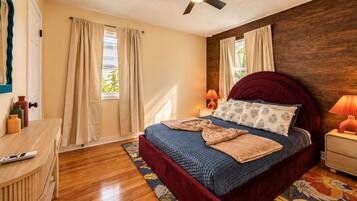 Une chambre à coucher comprenant un lit, un ventilateur de plafond, une table de chevet, une lampe, une commode en bois et une fenêtre avec des rideaux.