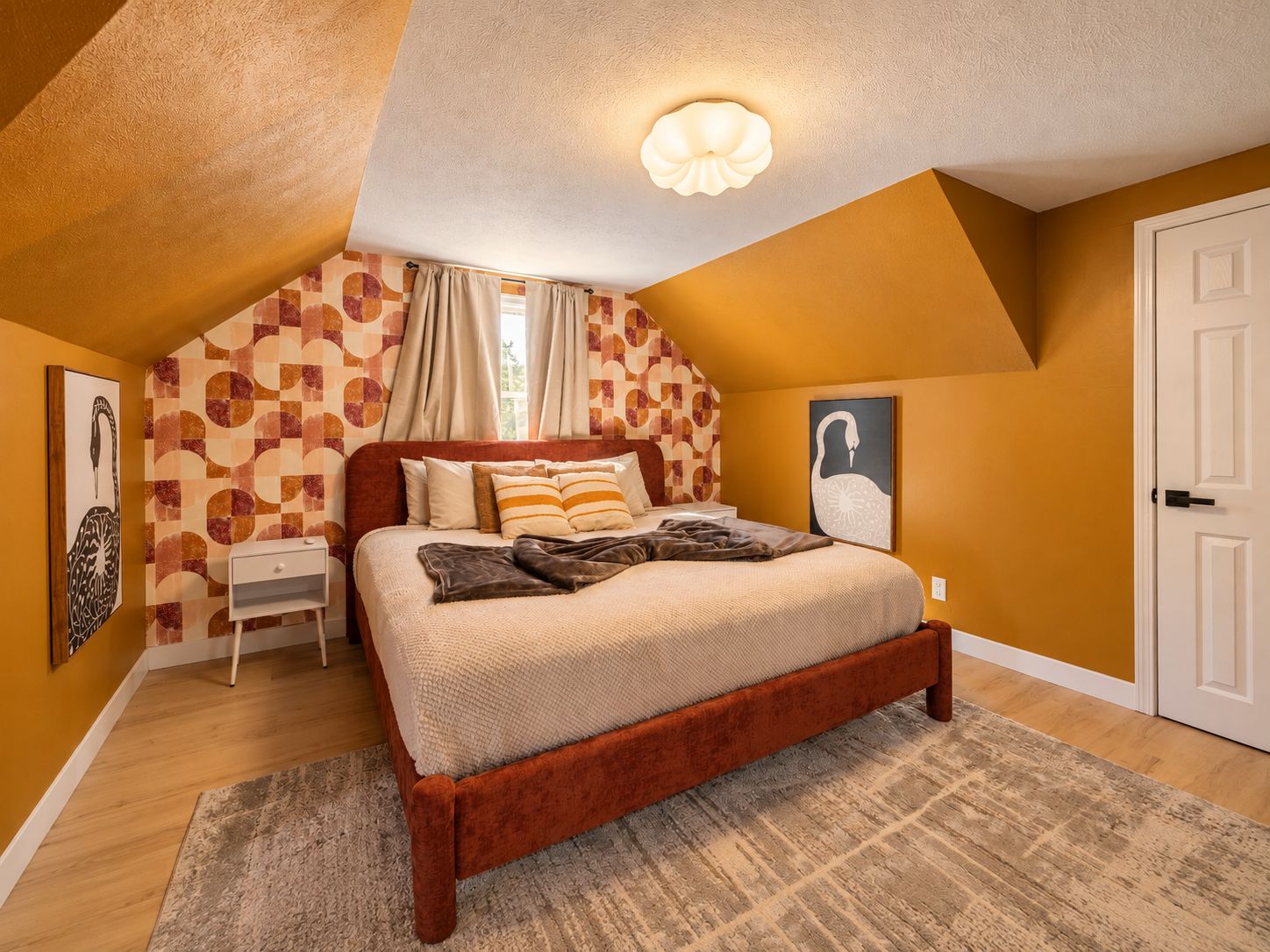 Una camera da letto con soffitto spiovente, un letto con struttura in legno, un comodino, una parete con carta da parati a motivi geometrici e una porta.