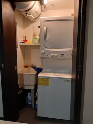 A compact laundry room with a front-loading washing machine, a small sink, and a shelf with cleaning supplies. - Nunciatura King Bed | Pool + Parking | Ü Tower, San José, Costa Rica. (San José)