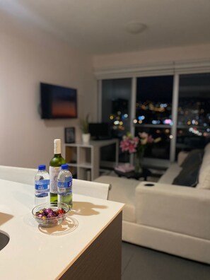 Ein modernes Wohnzimmer mit Essbereich, mit einem weißen Sofa, einem Glastisch mit Wasserflaschen und einer Schüssel Süßigkeiten, und Blick auf die Stadt durch große Fenster.