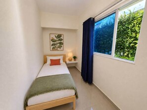 Ein Einzelbett mit Kopfteil, ein Nachttisch mit Lampe, ein Fenster mit Vorhängen und eine Wandpflanze.