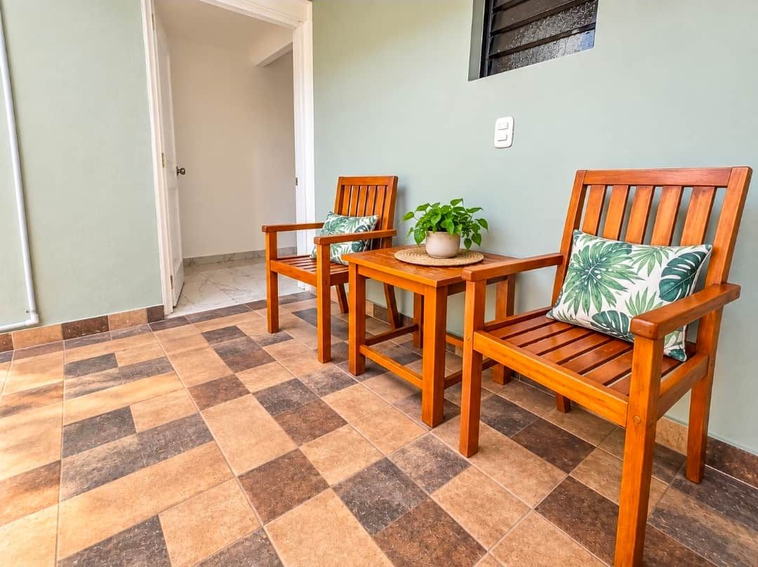 Una habitación con piso a cuadros, dos sillas de madera con cojines, una mesita con planta en maceta y un interruptor de luz en la pared.