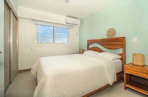 Un dormitorio con cama, mesita de noche, aire acondicionado instalado en la pared y ventana con vista al cielo.