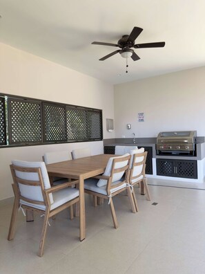 A dining area with a wooden table and chairs, a ceiling fan, and a built-in grill station. - Live Jaco 1207 Ocean View 2BR Condo Near Beach (Jaco)