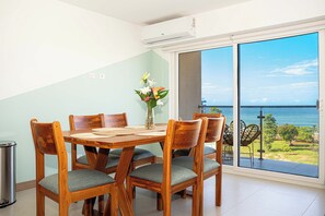 Un coin repas avec une table et des chaises en bois, un vase rempli de fleurs et une vue sur la mer, visible à travers une porte-fenêtre coulissante.