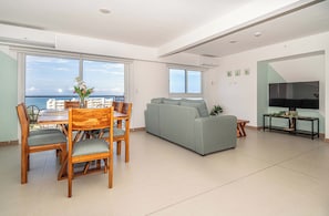 A modern living room with a dining area, a grey sofa, and a TV on a stand. - Live Jaco 1207 Ocean View 2BR Condo Near Beach (Jaco)