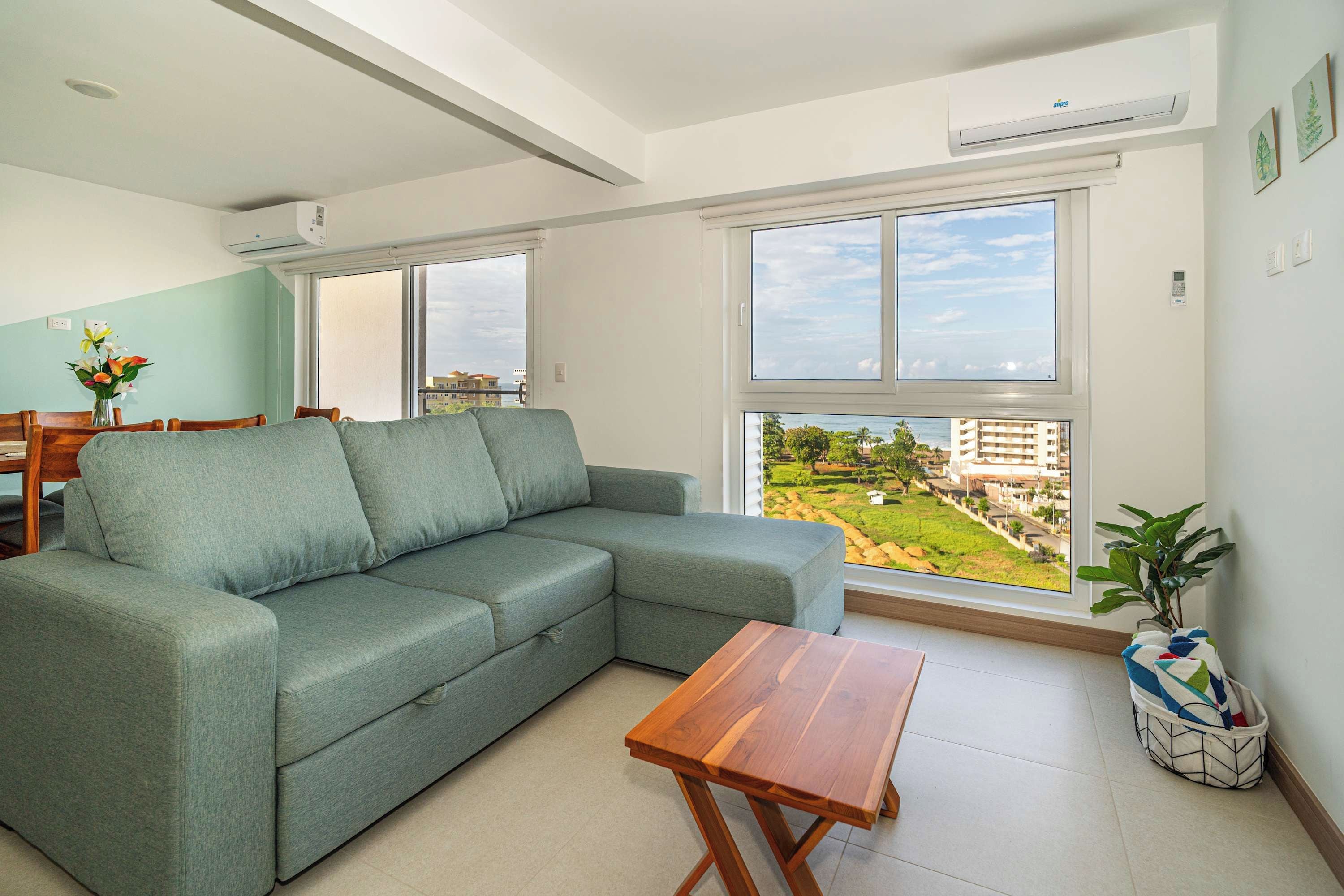 A modern living room with a sectional sofa, a wooden coffee table, and a view of a residential area through large windows.