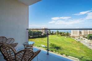 Un balcon agrémenté de meubles en osier, d’une table en verre et offrant une vue sur un parc verdoyant, des immeubles et la mer.