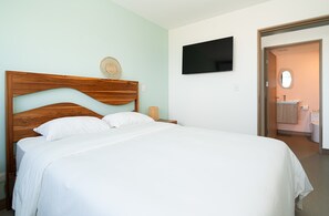 Un lit bien fait, avec une tête de lit en bois, un téléviseur à écran plat fixé au mur et une salle de bain visible à travers une porte ouverte.