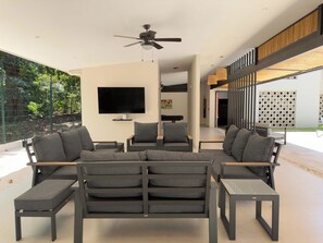 A modern living room with a sectional sofa, a flat-screen TV, and a ceiling fan. - Live Jaco 1207 Ocean View 2BR Condo Near Beach (Jaco)