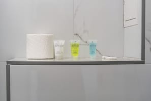 A bathroom counter with toiletries including a large roll of toilet paper, a hand and body lotion, a shampoo and conditioner, and a bath and shower gel. - Sutherland Apartments - Apt 26 (London)