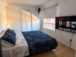 Une chambre à coucher avec un lit, un téléviseur fixé au mur affichant l’interface de Netflix, une fenêtre donnant sur un immeuble en briques et une porte.