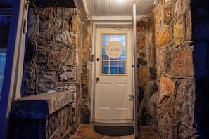 Un couloir aux murs de pierre mène à une porte sur laquelle est apposée une plaque indiquant : « The Riverstone House ».