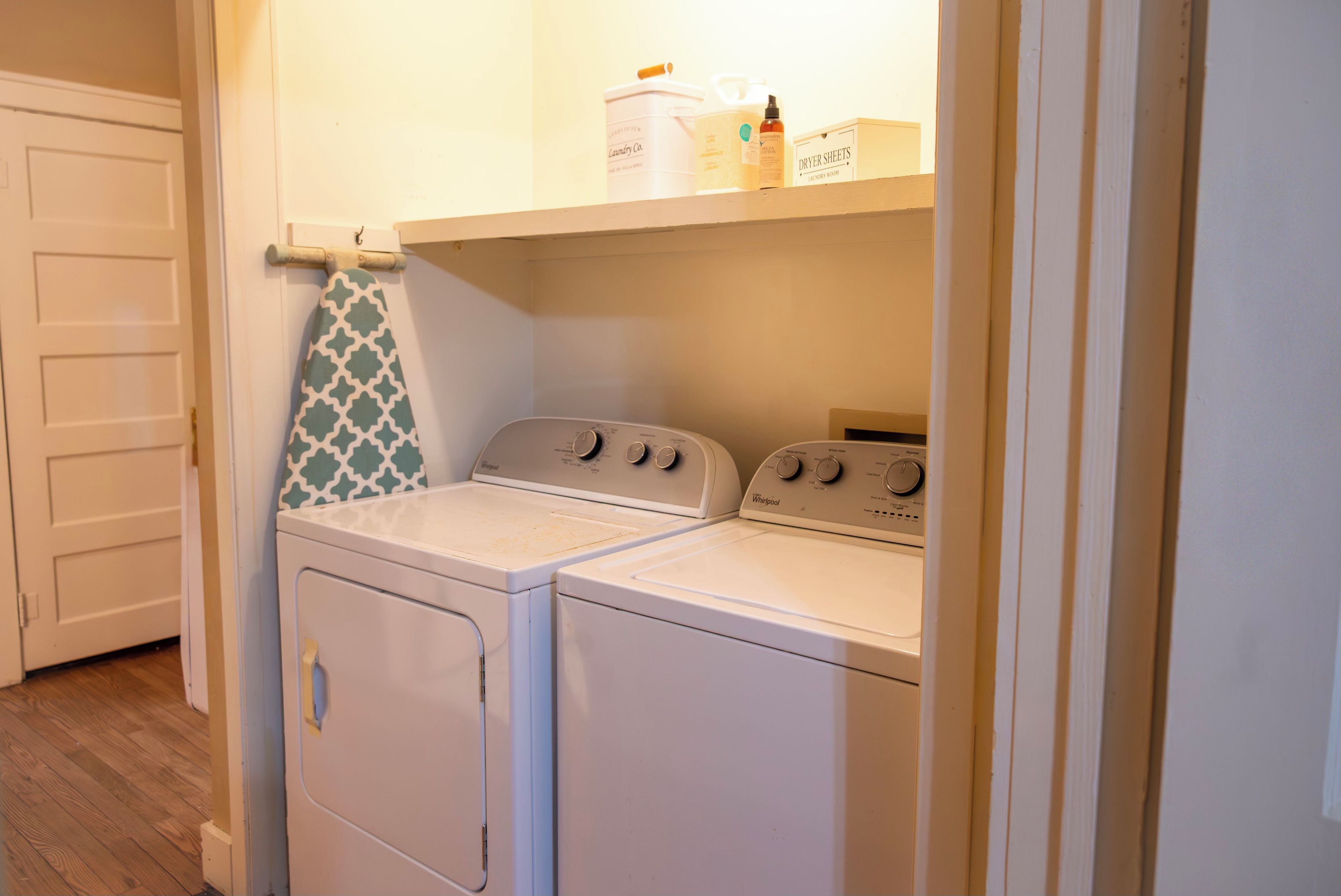 Une buanderie équipée de deux machines à laver et de deux sèche-linge Whirlpool blancs, d’un étendoir sur lequel est suspendue une serviette à motifs, et d’étagères contenant des produits de lessive.