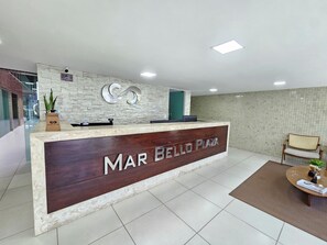 A reception desk with a sign reading 'MAR BELLO PLAZA' in a modern lobby with a tiled floor and a seating area with a round table. - MicroEstudio Intermares - 100m from the sea + cost-benefit (Cabedelo)
