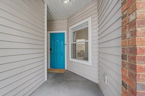 Ein schmaler Gang mit einer blauen Tür und einem Fenster, der zu einem Zimmer mit einem sichtbaren Badezimmer führt.