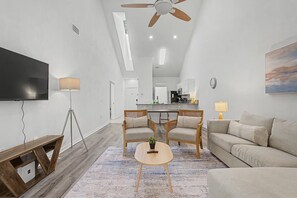 Ein modernes Wohnzimmer mit Flachbildfernseher, Deckenventilator, Essbereich und einer Küche im Hintergrund sowie einem großen Fenster.