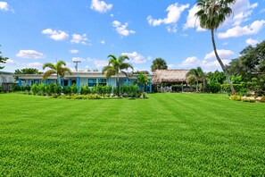 A well-maintained lawn with a building featuring blue and white exterior walls, palm trees, and a thatched-roof structure. - The Conchy Cabana- Unit 2 (Jensen Beach)