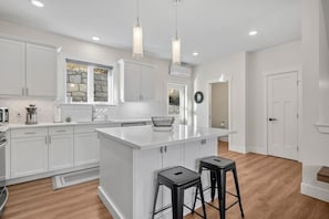 A modern kitchen with white cabinetry, a central island, two barstools, pendant lighting, and a stone backsplash. - Cherry Cove | Beach Access | AC | BBQ | Dbl Garage (Invermere)