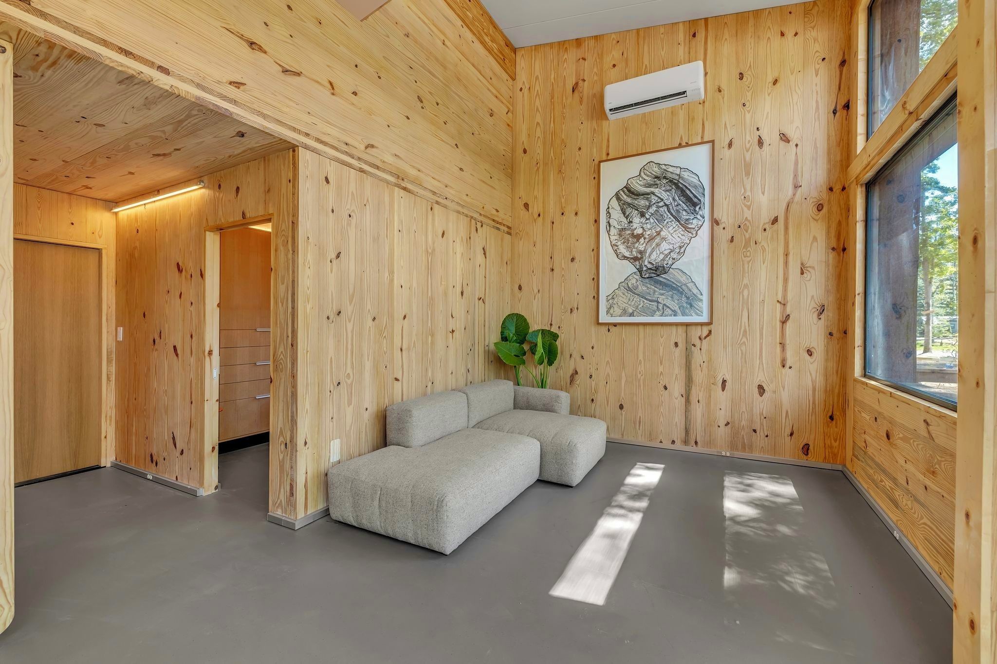 Une pièce aux murs en bois, avec un canapé gris, une œuvre d’art encadrée et une fenêtre.