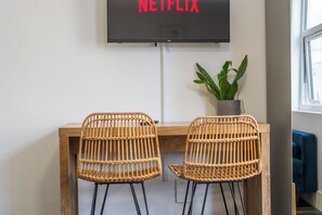 A room with a wooden desk, two wicker chairs, a potted plant, and a TV mounted on the wall displaying 'NETFLIX'. - Sutherland Apartments - Apt 27 (London)