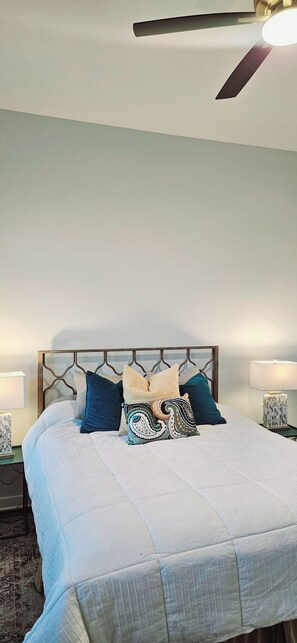 A neatly made bed with a patterned bedspread and multiple pillows, a bedside table with a lamp, and a ceiling fan. - Contemporary two-story home close to the American Museum of Fine Art (Little Rock)