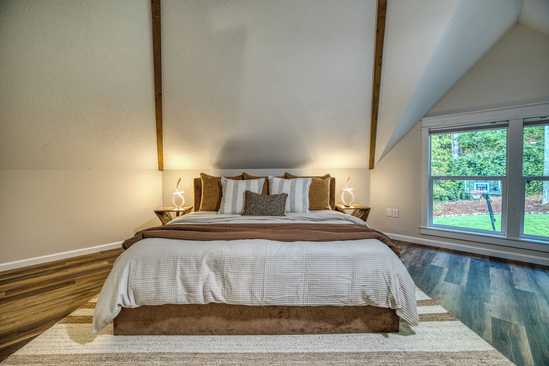 A bedroom with a large bed, two bedside tables, a window with a view of greenery, and a sloped ceiling.