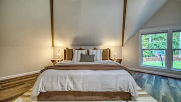 A bedroom with a large bed, two bedside tables, a window with a view of greenery, and a sloped ceiling.