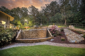 A landscaped garden with a stone water feature, a wooden deck, and a well-maintained lawn. - Private Pool, Fenced Yard, Sauna, Hot Tub & Outdoor Fireplace | 5 Min downtown (Ellijay)