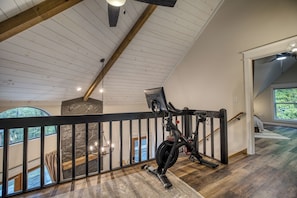 A spacious room with a sloped ceiling, a stationary exercise bike, and a view of the outdoors through a large window. - Private Pool, Fenced Yard, Sauna, Hot Tub & Outdoor Fireplace | 5 Min downtown (Ellijay)