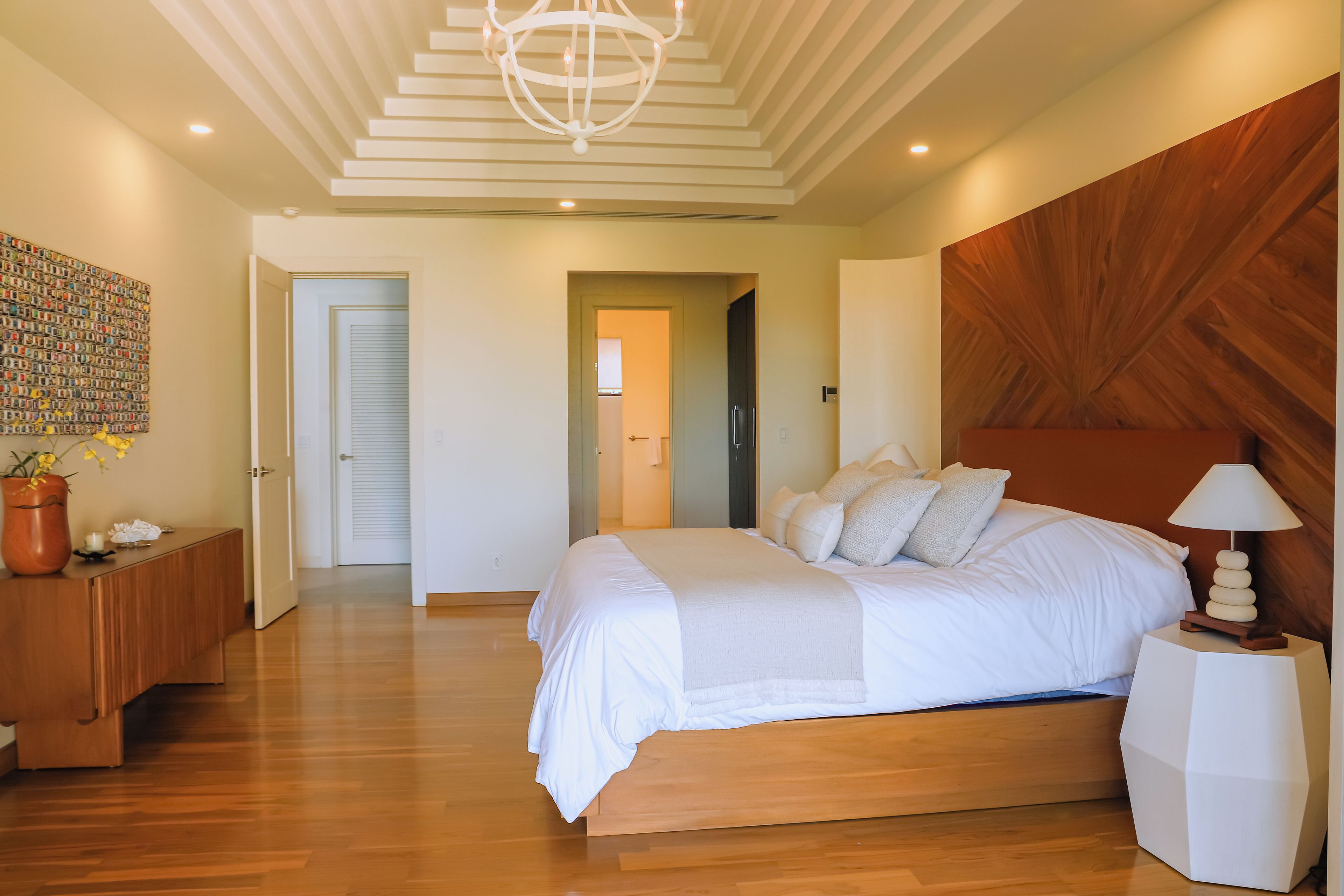 A modern bedroom with a large bed, wooden flooring, a chandelier, and a unique wooden wall panel.