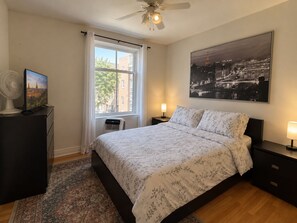 Une chambre à coucher comprenant un lit, une table de chevet, une lampe, un ventilateur, une fenêtre et un tableau représentant un paysage urbain.