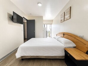 Une chambre à coucher avec un lit, une table de chevet en bois, un téléviseur fixé au mur et une porte donnant sur un balcon.
