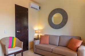 A room with a dark wooden door, a wall-mounted air conditioner, a round mirror, a woven chair with a cushion, a grey sofa with cushions, and a small table with a lamp. - Group Villa. Everyone Gets Their Own Suite. Private Pool. Heart of Sayulita. (Sayulita)