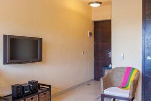 Habitación con una televisión de pantalla plana montada en la pared, un mueble negro para entretenimiento con altavoces, un sillón de mimbre con una manta colorida y una puerta con picaporte.