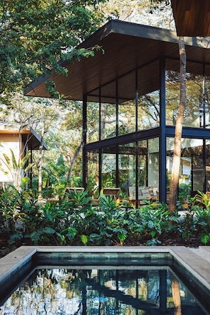 A modern outdoor structure with a glass enclosure, surrounded by lush greenery and a reflective pool.