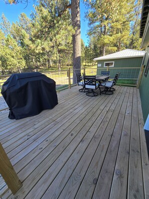 Eine Holzterrasse mit Grill, Gartentisch und Stühlen sowie ein grünes Gebäude im Hintergrund.