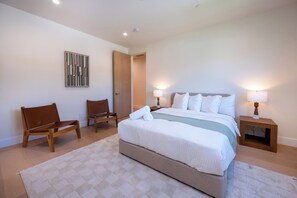 A bedroom with a bed, two chairs, a bedside table with a lamp, a door, and a wall-mounted artwork. - Gaviota Pavilion By Alora Reserve (Los Angeles)