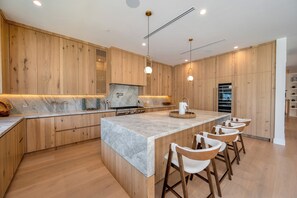 A modern kitchen with wooden cabinetry, a marble countertop, and bar stools. - Gaviota Pavilion By Alora Reserve (Los Angeles)