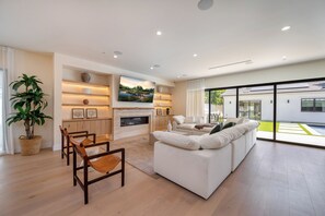 A modern living room with a fireplace, a sofa set, a dining area, and a large window offering a view of the outdoors. - Gaviota Pavilion By Alora Reserve (Los Angeles)