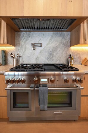 A kitchen with a stainless steel range hood, double ovens, and a marble backsplash. - Gaviota Pavilion By Alora Reserve (Los Angeles)