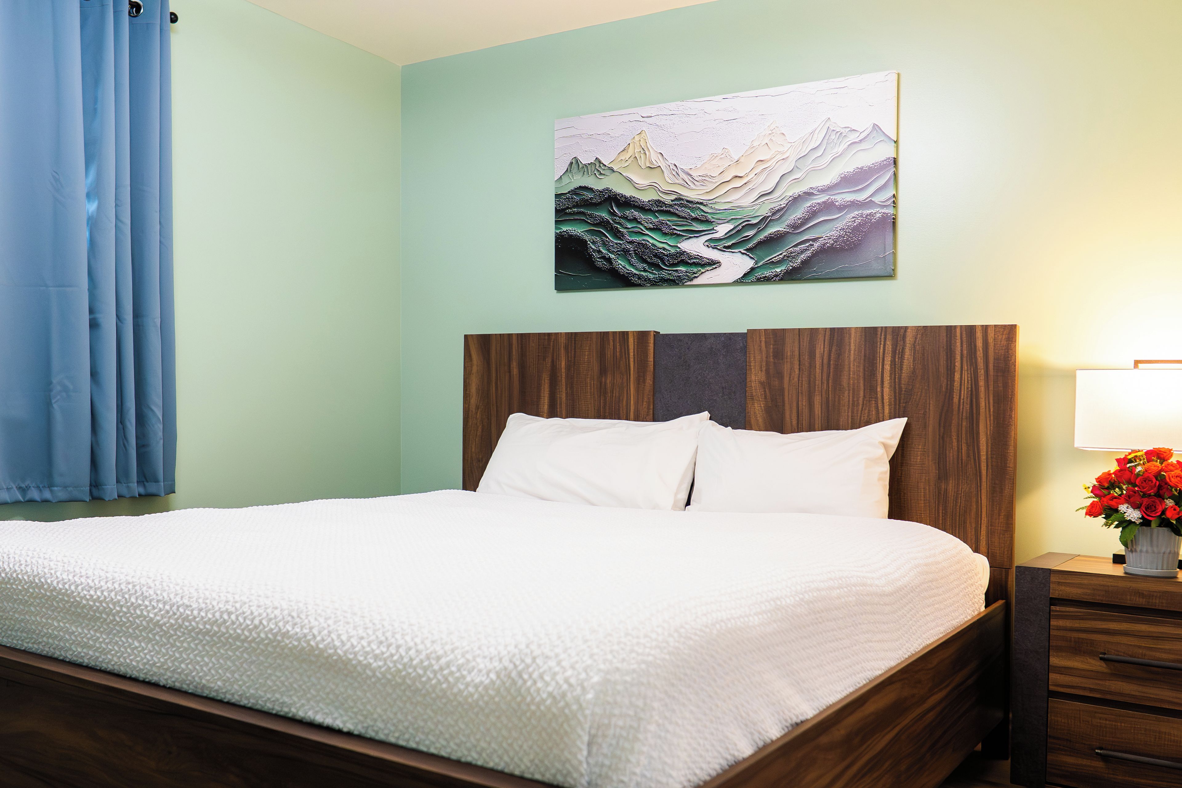 Un lit bien fait, avec une literie blanche, une tête de lit en bois et un tableau représentant un paysage de montagne accroché au mur.