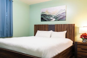 A neatly made bed with white bedding, a wooden headboard, and a wall-mounted painting of a mountain landscape. - Casa SKKY (Whitehorse)