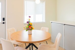 A round table with a white vase of flowers, surrounded by white chairs, in a room with a window and a closed cabinet. - Casa SKKY (Whitehorse)