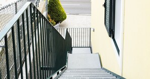 Eine Treppe führt zu einem Gebäudeeingang, links daneben ein Maschendrahtzaun und im Hintergrund ein kleiner Baum.