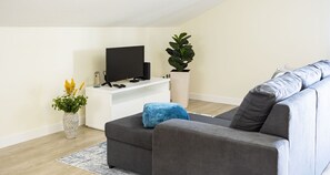 Ein modernes Wohnzimmer mit einem grauen Sofa, einem Flachbildfernseher auf einem weißen Standfuß, einer Topfpflanze und einer Vase mit Blumen.