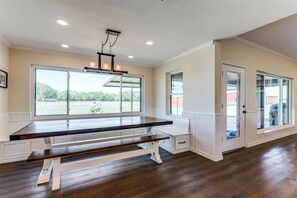A spacious room with a large dining table, built-in bench seating, and large windows offering a view of the outdoors.