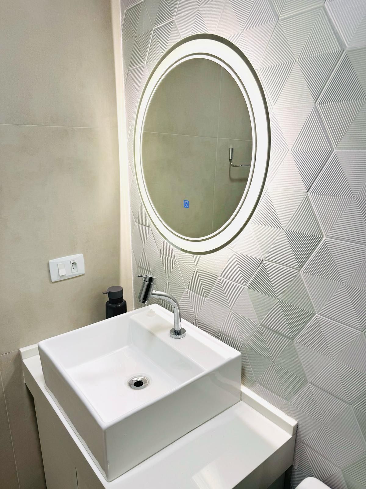 Ein Badezimmer mit einem weißen Waschbecken, einem runden Spiegel mit Beleuchtung und einer Wand mit geometrischem Muster.