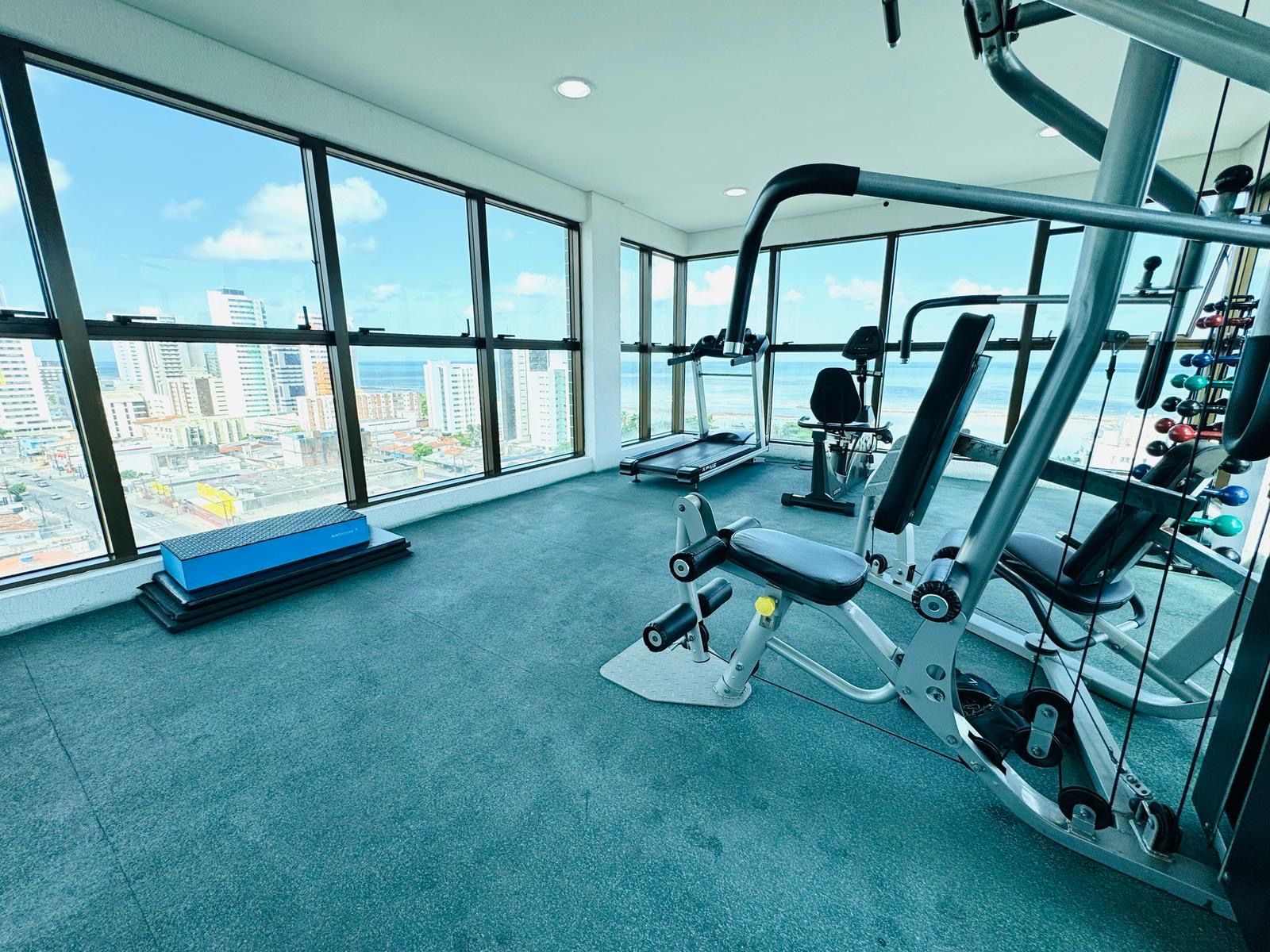 Un gimnasio con equipo de ejercicio, una banca y vistas a un paisaje urbano a través de amplias ventanas.